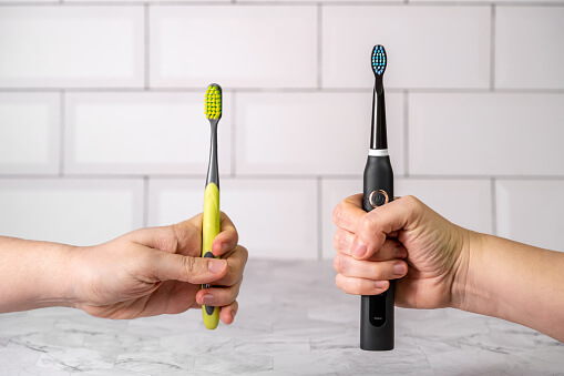 Two hands hold toothbrushes for comparison: one hand displays a yellow-handled manual toothbrush with green bristles, while the other showcases a black electric toothbrush with blue bristles. Background features white tiled walls and a marble-like surface.