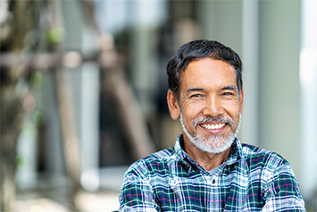 A smiling older man with a gray beard and wearing a plaid shirt stands outdoors