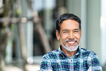 A smiling older man with a gray beard and wearing a plaid shirt stands outdoors