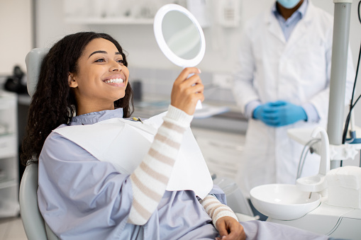 Happy Female Patient Looking At Mirror After Dental Treatment In Clinic Sitting In a dental Chair, enjoying Her Beautiful Smile.