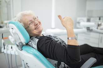 Older adult in turquoise dental chair smiles and gives thumbs‑up, dental equipment visible in clinic background.